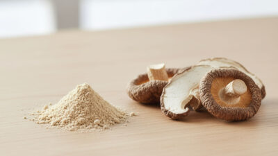 A small pile of beige mushroom powder sits next to several slices of dried shiitake mushrooms on a light wooden surface, with a blurred background.