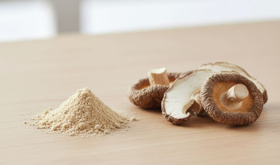A small pile of beige mushroom powder sits next to several slices of dried shiitake mushrooms on a light wooden surface, with a blurred background.