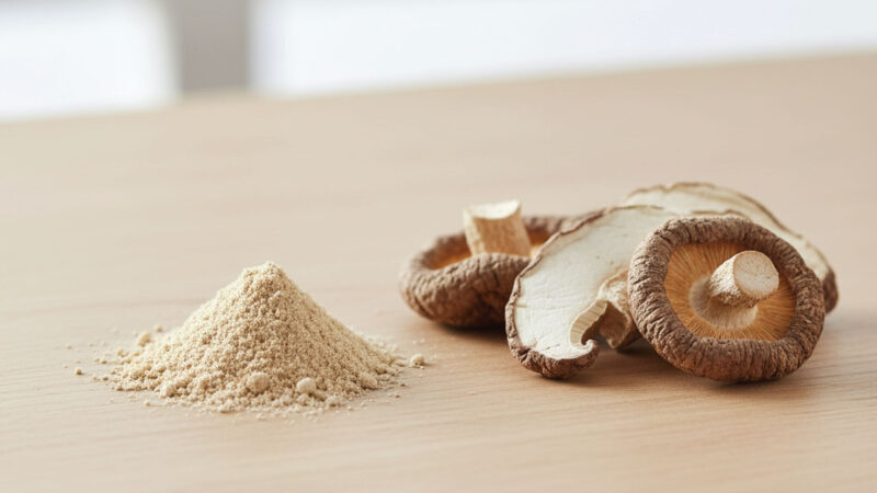 A small pile of beige mushroom powder sits next to several slices of dried shiitake mushrooms on a light wooden surface, with a blurred background.