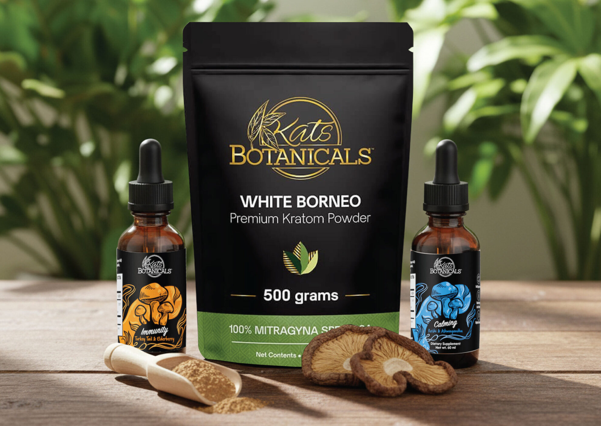 A black bag labeled Kats Botanicals White Borneo Premium Kratom Powder 500 grams sits on a wooden table, flanked by two dropper bottles and a wooden scoop of powder. Green plants are blurred in the background.