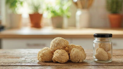 Several lions mane mushrooms are placed on a wooden table beside a glass jar containing two more mushrooms, with a kitchen and potted plants blurred in the background.