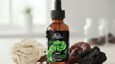 A brown glass dropper bottle labeled Kats Botanicals sits on a light surface, surrounded by different dried mushrooms in white, brown, and black hues. The bottle has a green label with mushroom illustrations.