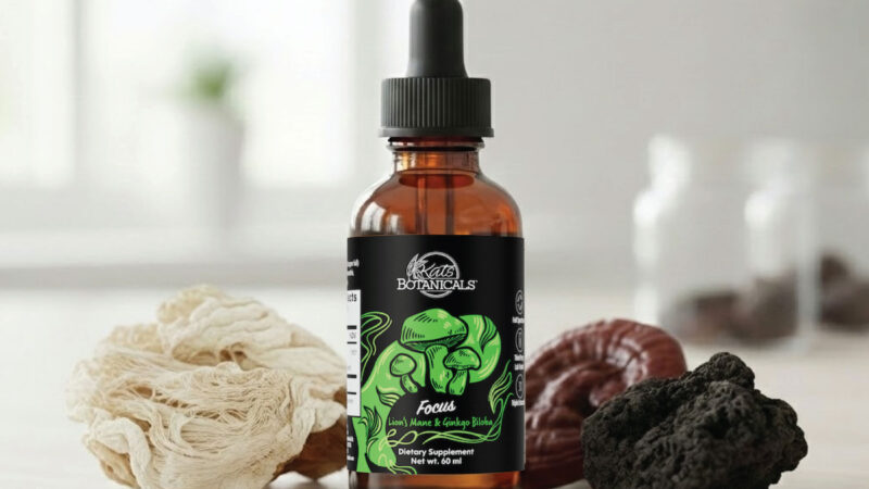 A brown glass dropper bottle labeled Kats Botanicals sits on a light surface, surrounded by different dried mushrooms in white, brown, and black hues. The bottle has a green label with mushroom illustrations.