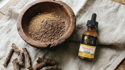 A wooden bowl filled with brown powder sits on a table next to a dropper bottle labeled “Katra Rava Premium Goat Tincture.” Dried root pieces are also on the table beside the bowl.