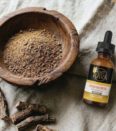A wooden bowl filled with brown powder sits on a table next to a dropper bottle labeled “Katra Rava Premium Goat Tincture.” Dried root pieces are also on the table beside the bowl.