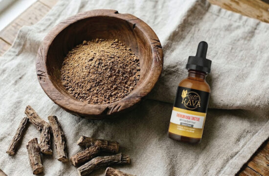 A wooden bowl filled with brown powder sits on a table next to a dropper bottle labeled “Katra Rava Premium Goat Tincture.” Dried root pieces are also on the table beside the bowl.