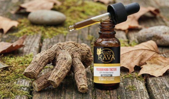 A bottle labeled Kats Kava Premium Kava Tincture stands on a wooden surface with its dropper resting on top. Next to it are kava roots, moss, fallen leaves, and smooth stones in the background.