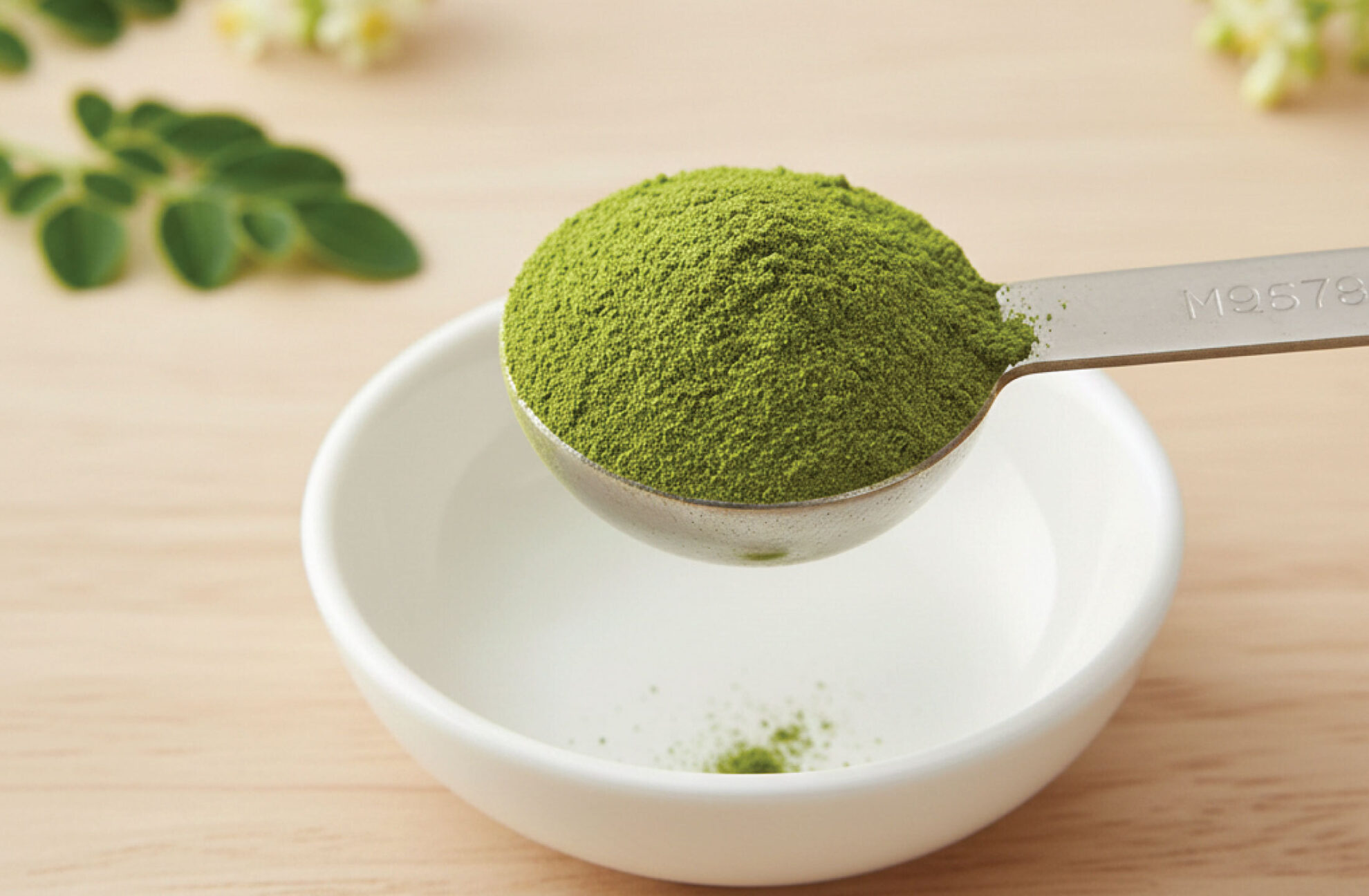  A metal tablespoon holds vibrant green powder above a small white bowl on a light wooden surface, with a few green leaves blurred in the background.