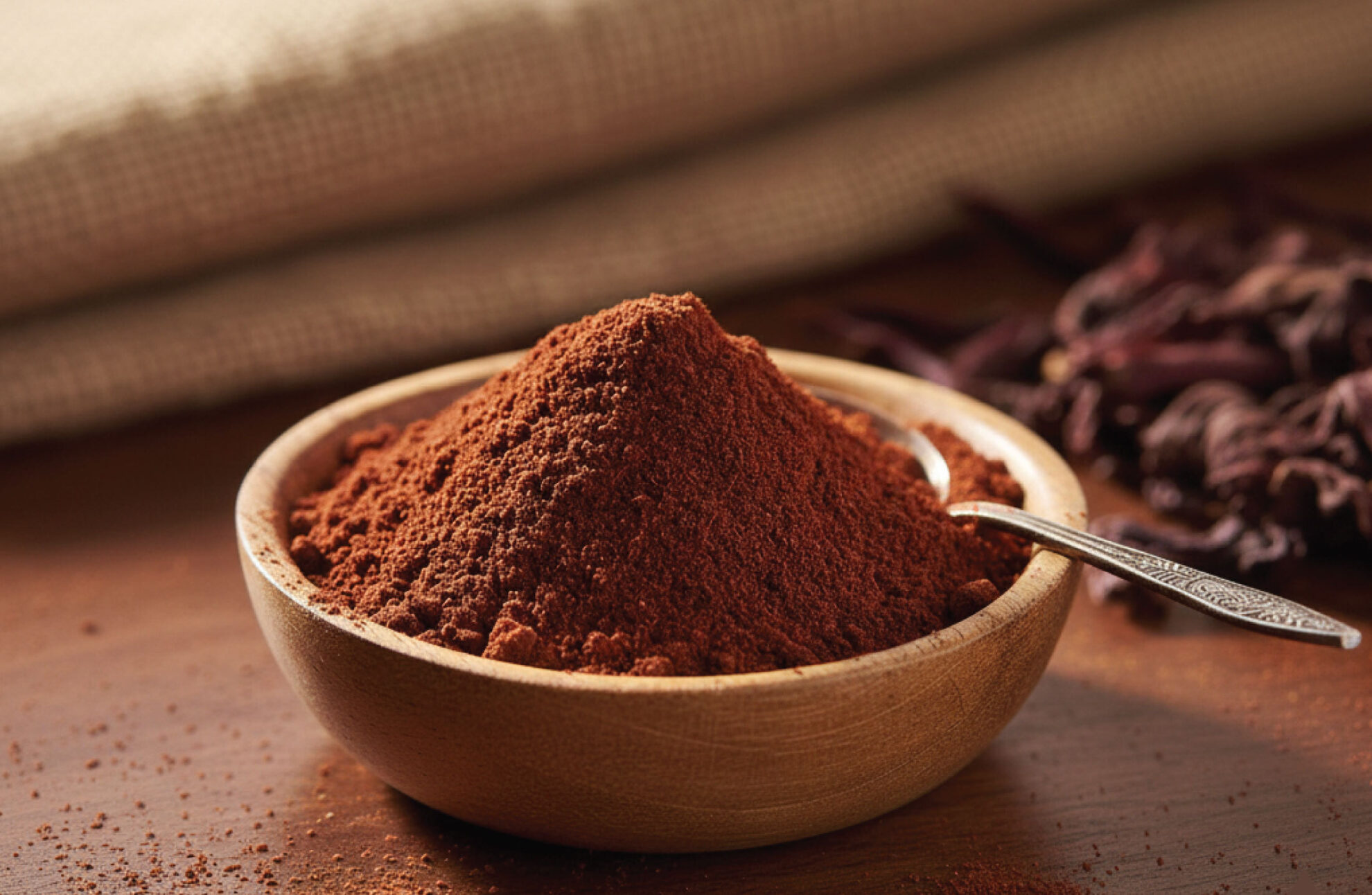 A wooden bowl filled with reddish-brown red maeng da kratom powder sits on a wooden surface. A silver spoon rests in the bowl, and red maeng da kratom is partially visible in the background.