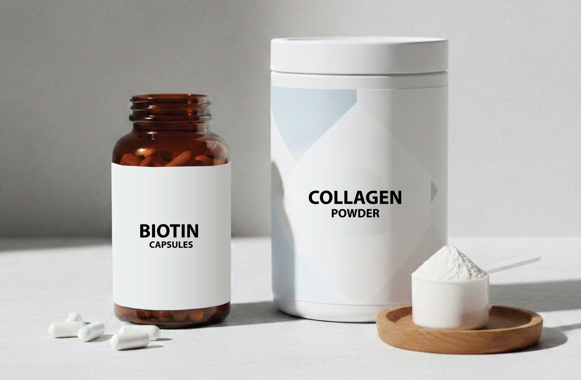 A brown bottle labeled Biotin Capsules is next to a white container labeled Collagen Powder with a small wooden tray holding a scoop of white powder and some capsules on a light surface.
