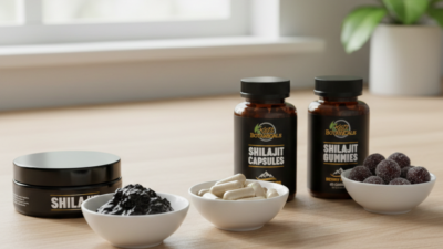 Three small white bowls hold shilajit resin, capsules, and gummies in front of labeled jars for shilajit resin, capsules, and gummies on a light wooden table with a blurred window in the background.