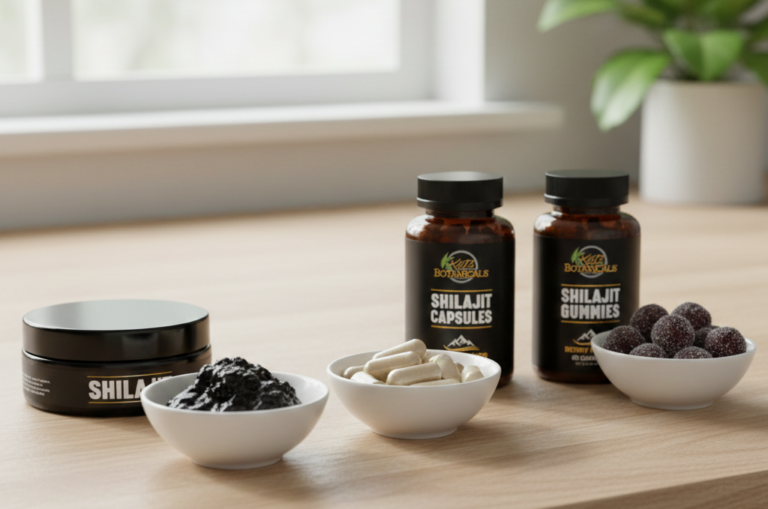 Three small white bowls hold shilajit resin, capsules, and gummies in front of labeled jars for shilajit resin, capsules, and gummies on a light wooden table with a blurred window in the background.