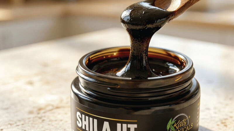 A wooden spoon lifts thick, dark shilajit resin from an open jar labeled SHILAJIT by Kats Botanicals, resting on a sunlit kitchen counter.