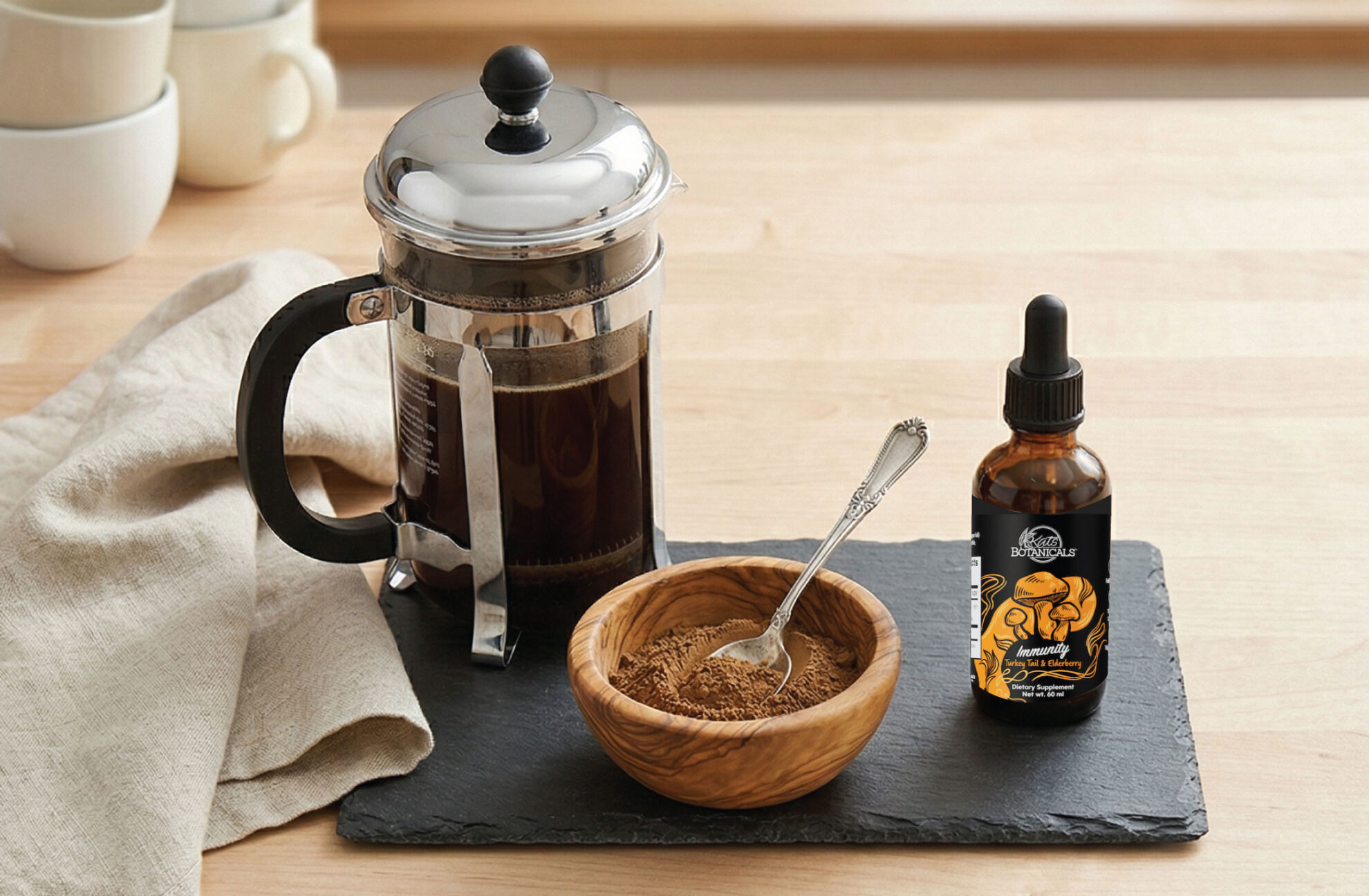 A French press with brewed coffee, a wooden bowl of powder with a spoon, and a dropper bottle are arranged on a black slate tray atop a wooden table, with stacked white cups in the background.
