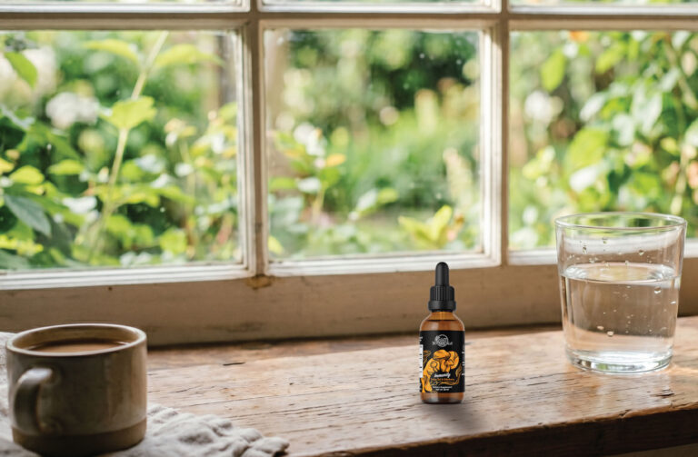 A small dark bottle with a dropper, a glass of water, and a ceramic mug sit on a wooden windowsill with a green, leafy garden visible through the window.