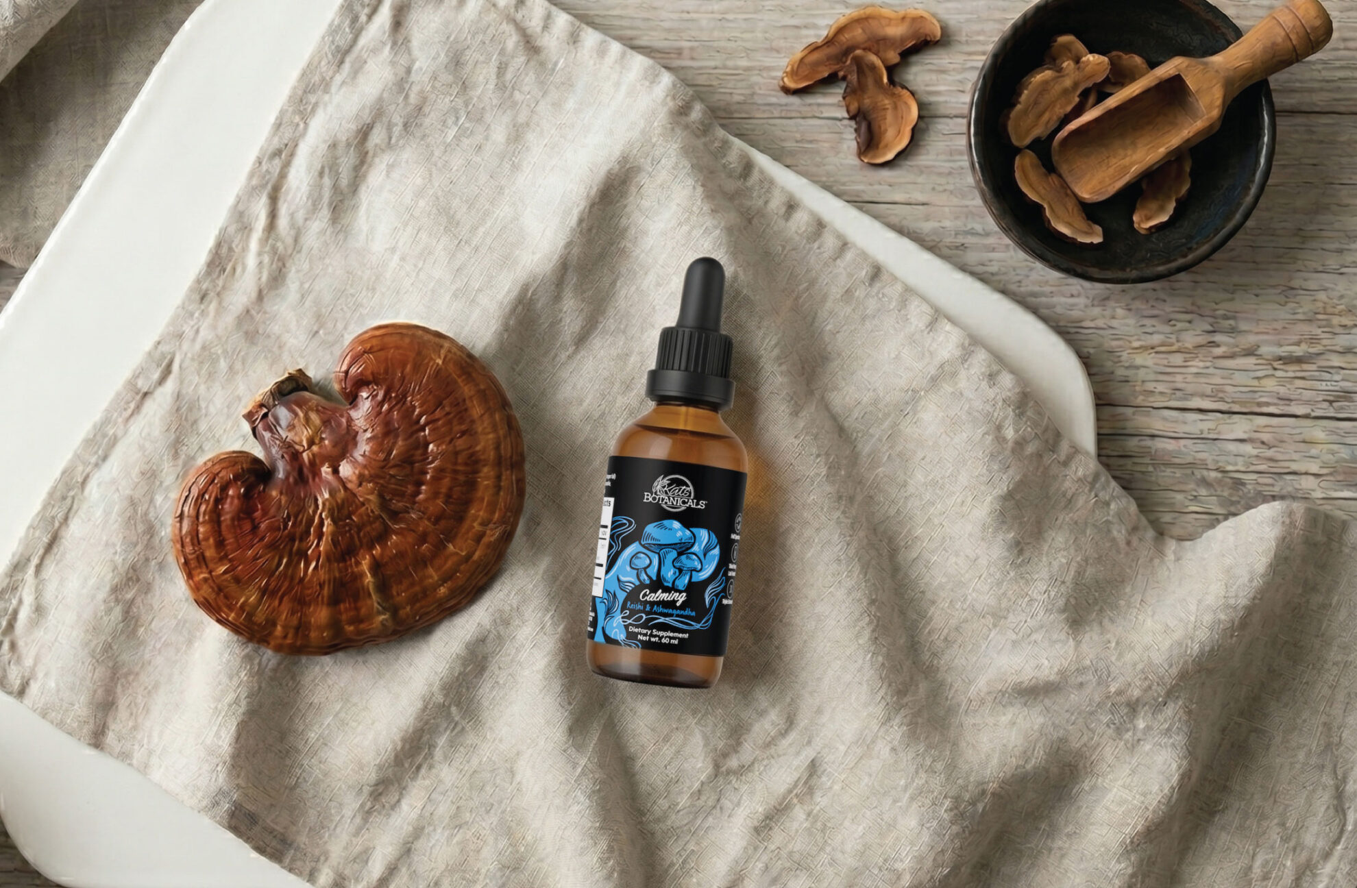 A brown glass dropper bottle with a blue label sits on beige fabric next to a large reishi mushroom, wooden bowl, and mushroom slices on a light wooden surface.