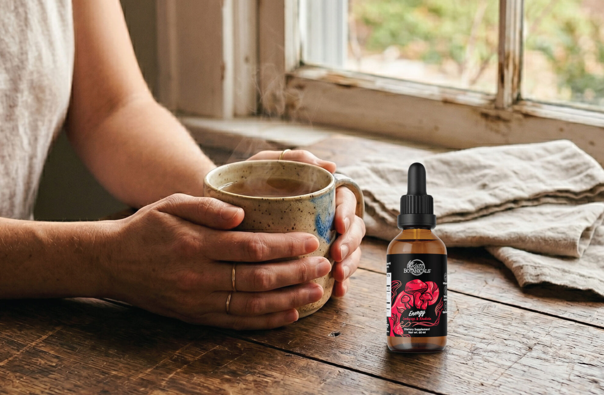 A person holds a steaming mug at a wooden table near a window, with a dropper bottle labeled Energy featuring a red illustration placed on the table beside them.