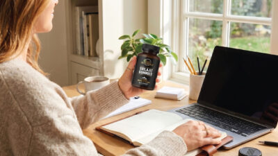 A person sits at a desk with an open notebook, laptop, and pens, holding a bottle labeled “Shilajit Gummies” near a window with sunlight streaming in.