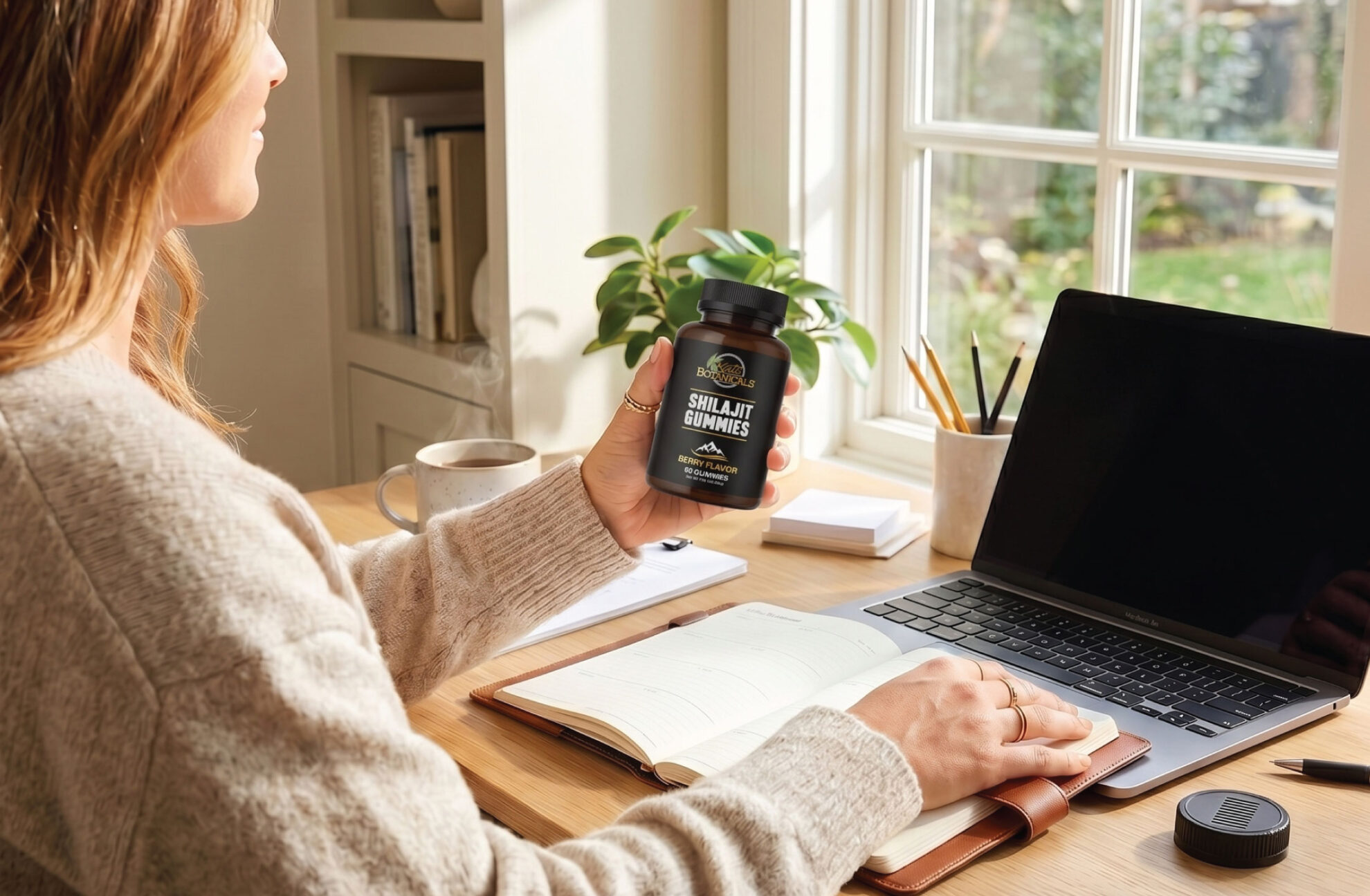 A person sits at a desk with an open notebook, laptop, and pens, holding a bottle labeled “Shilajit Gummies” near a window with sunlight streaming in.