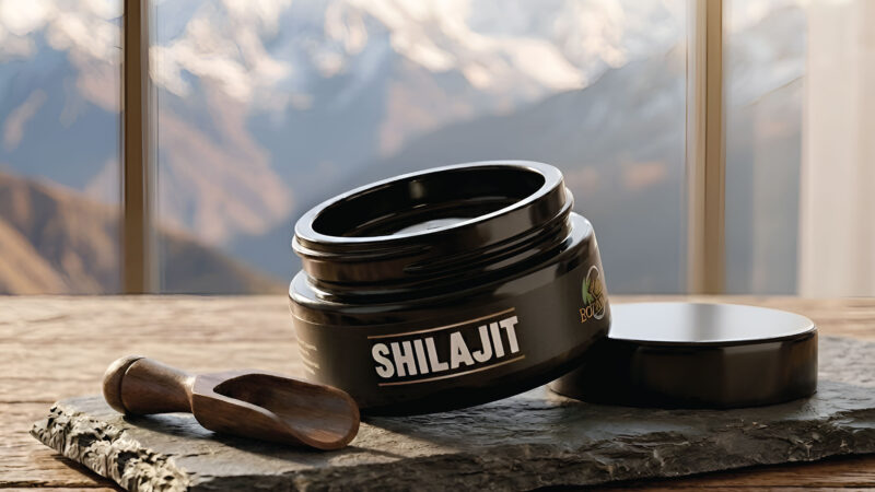 A black jar labeled Shilajit sits open on a stone slab with a small wooden spoon beside it; the background shows blurred mountains viewed through large windows.