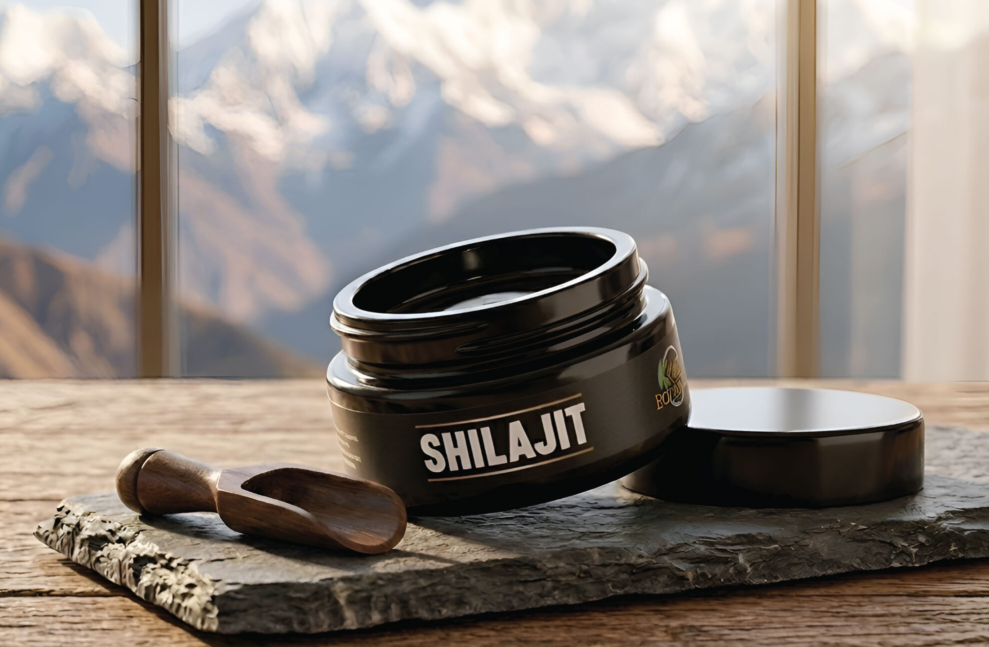 A black jar labeled Shilajit sits open on a stone slab with a small wooden spoon beside it; the background shows blurred mountains viewed through large windows.