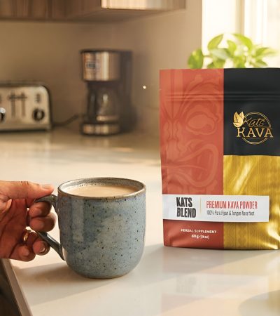 A hand holds a mug of kava in a bright kitchen, with a bag of Kat’s Kava premium kava powder on the counter beside a spoon, coffee maker, and toaster. Sunlight shines through the window.