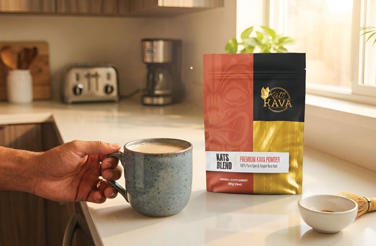 A hand holds a mug of kava in a bright kitchen, with a bag of Kat’s Kava premium kava powder on the counter beside a spoon, coffee maker, and toaster. Sunlight shines through the window.
