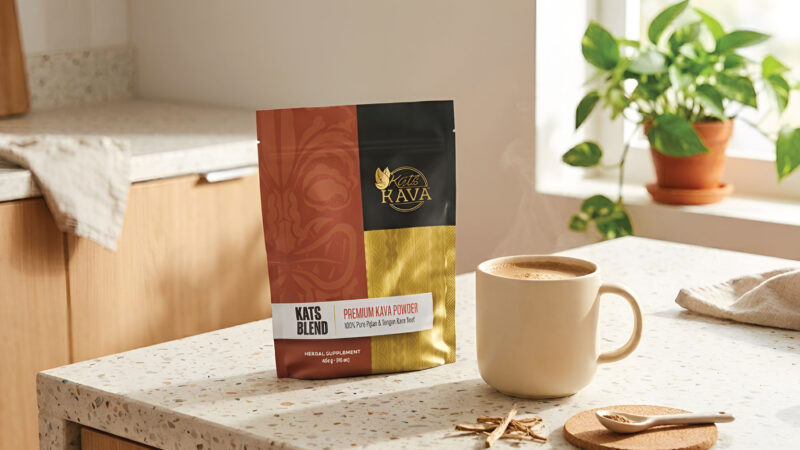 A bag of Kats Kava Kats Blend Premium Kava Powder stands on a kitchen counter next to a steaming mug, with a plant and kitchen cabinets in the background.