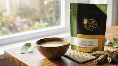 A wooden bowl filled with a light brown liquid sits next to a Kats Kava extract package on a wooden table, with sunlight streaming through a nearby window.