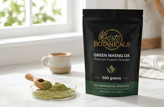 A bag of Kats Botanicals Green Maeng Da Kratom powder sits on a kitchen counter next to a mug, a cloth, and a small plate with powder and a wooden scoop.