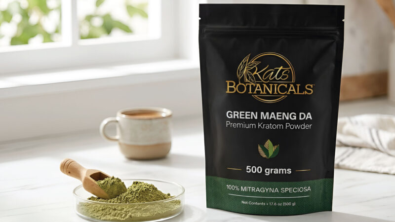 A bag of Kats Botanicals Green Maeng Da Kratom powder sits on a kitchen counter next to a mug, a cloth, and a small plate with powder and a wooden scoop.