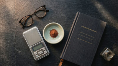 A digital scale, a pair of eyeglasses, a bowl with brown powder, a black notebook, and an ink bottle are arranged on a dark slate surface.