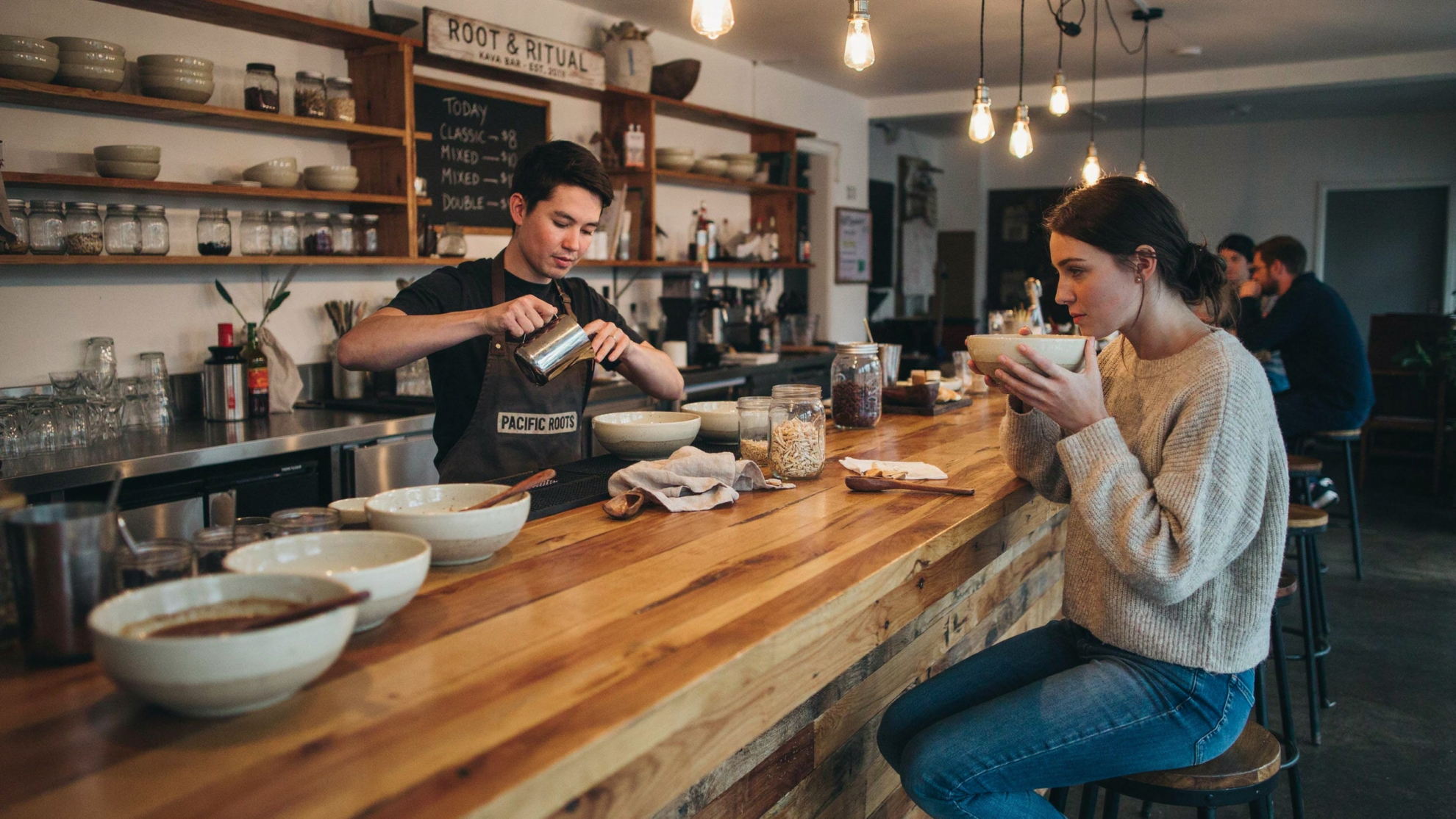 A barista in an apron pours liquid into a bowl for a woman sitting at a wooden counter in a cafe.